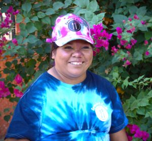 A lovely, young woman wearing a blue tye-dye shirt with a pink tye-dye golf cap. She is in front of pink flowers budding on a tree and has a wonderful smile.